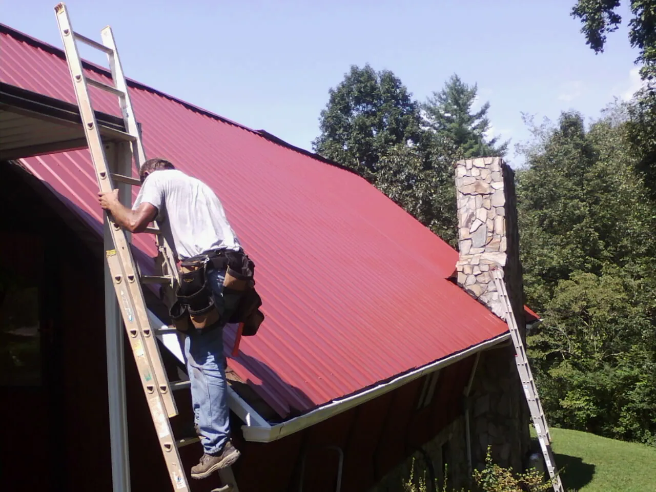 Roofer on a ladder beside a red metal roof and stone chimney — Soffit & Fascia Repair by Roofing Brothers in Clive